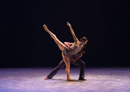 Two BalletX dancers performing a dynamic duet on stage. One dancer extends in an elegant arabesque while the other supports them with a grounded stance. They are dressed in contemporary costumes, and dramatic lighting highlights their fluid movement and expression.